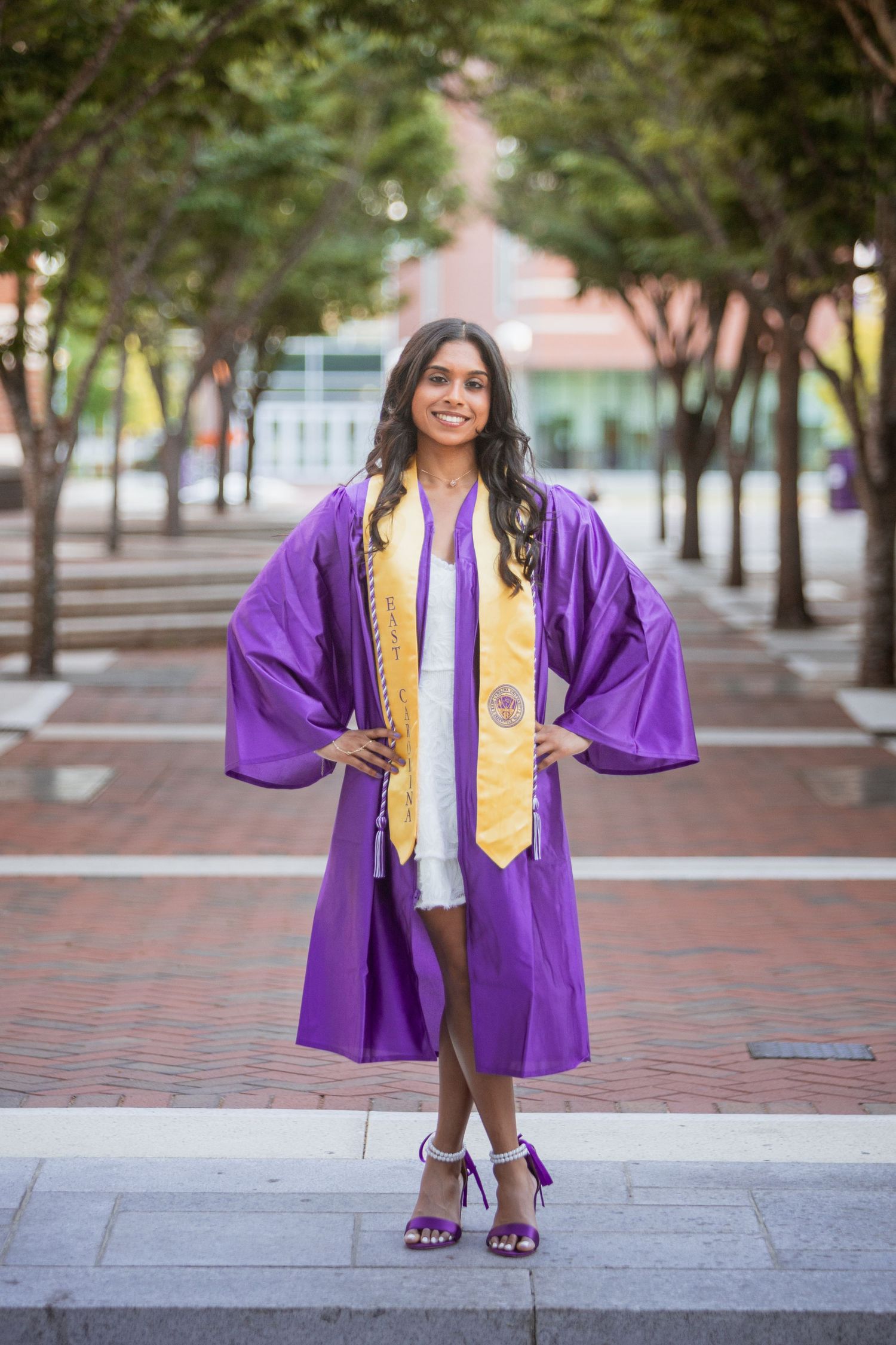 Young woman in purple graduation cap and gown posing on a brick walkway outdoors.