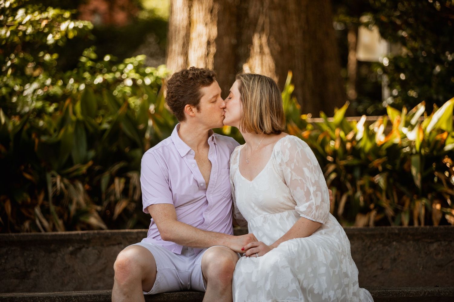 Couple sharing a romantic kiss while seated on stone steps surrounded by lush greenery.