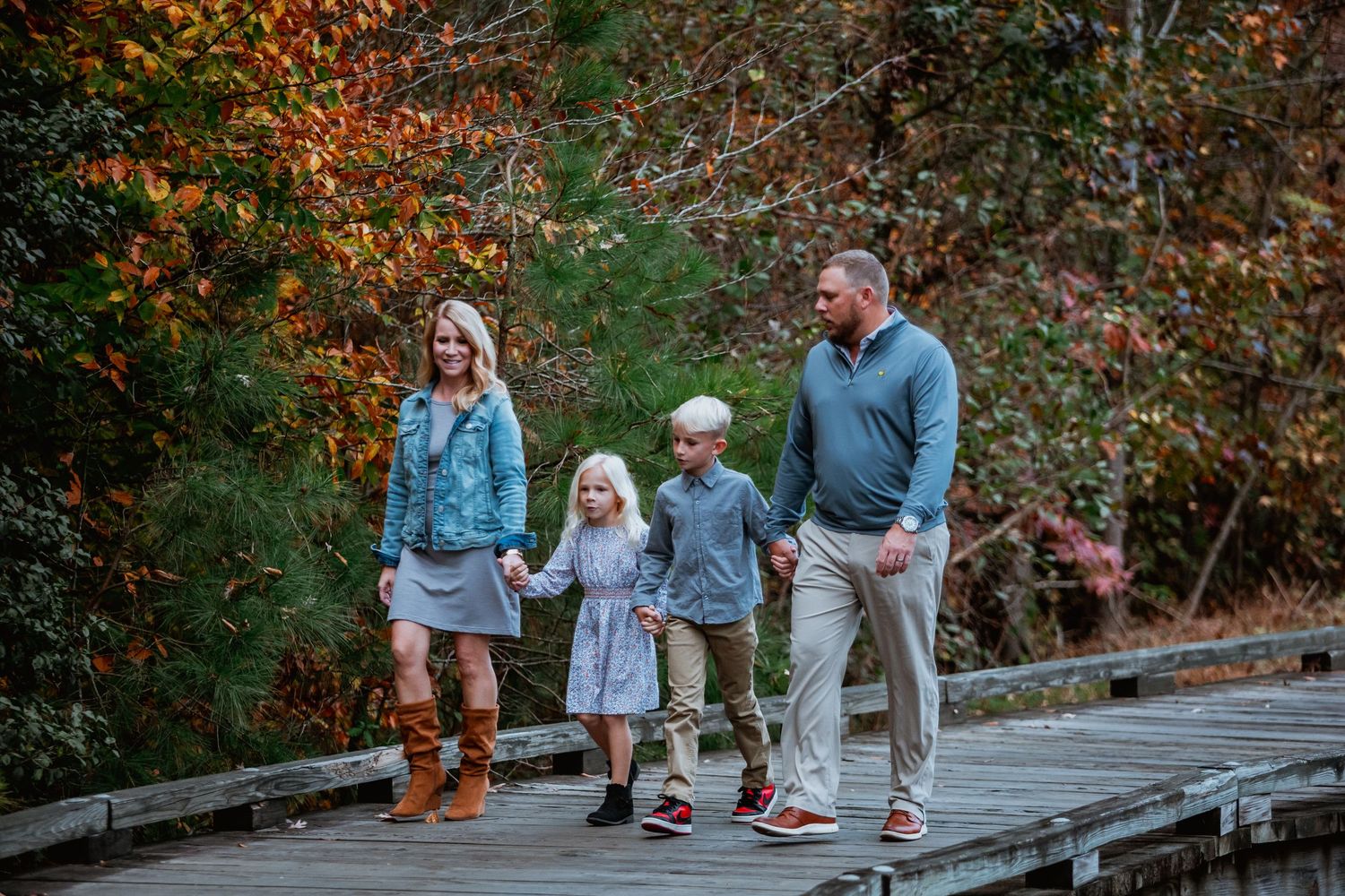 Family of four holding hands while walking on wooden bridge surrounded by autumn foliage.