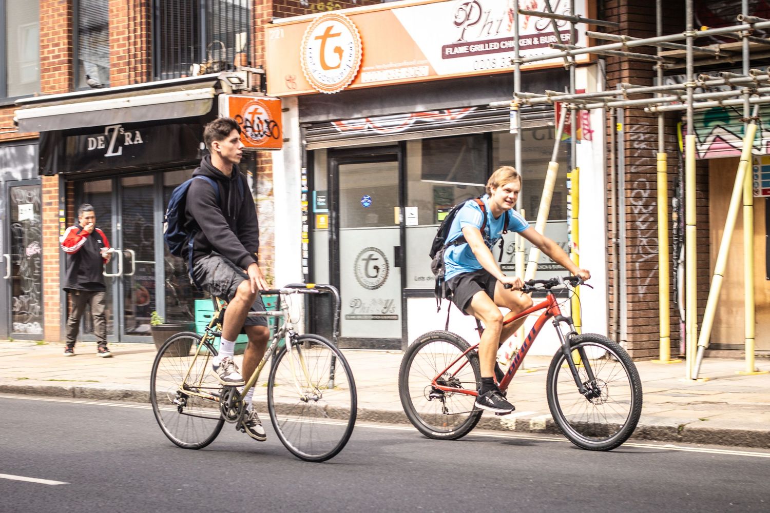 Two cyclists ride bicycles along a busy urban street past storefronts on a sunny day.