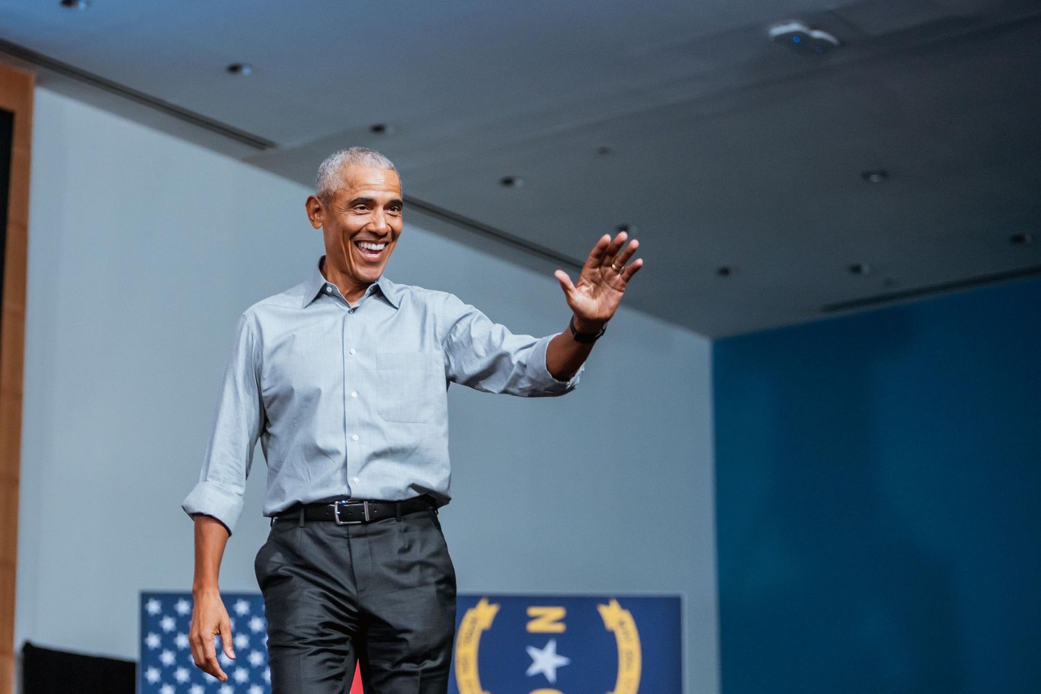 A man in a light blue shirt waves to a crowd on a decorated stage.