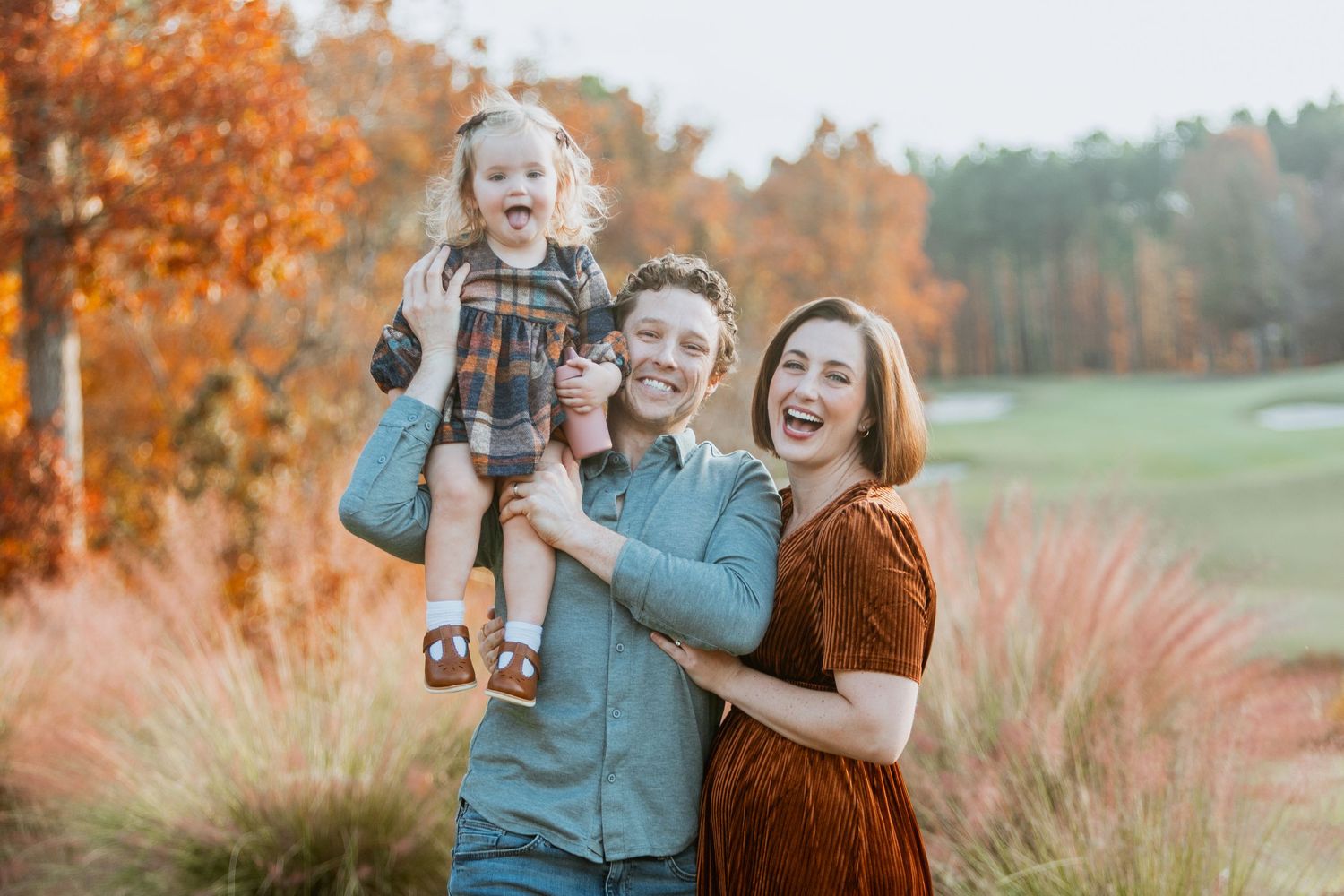 Joyful family of three laughs together as dad holds toddler daughter among vibrant orange fall foliage on golf course.