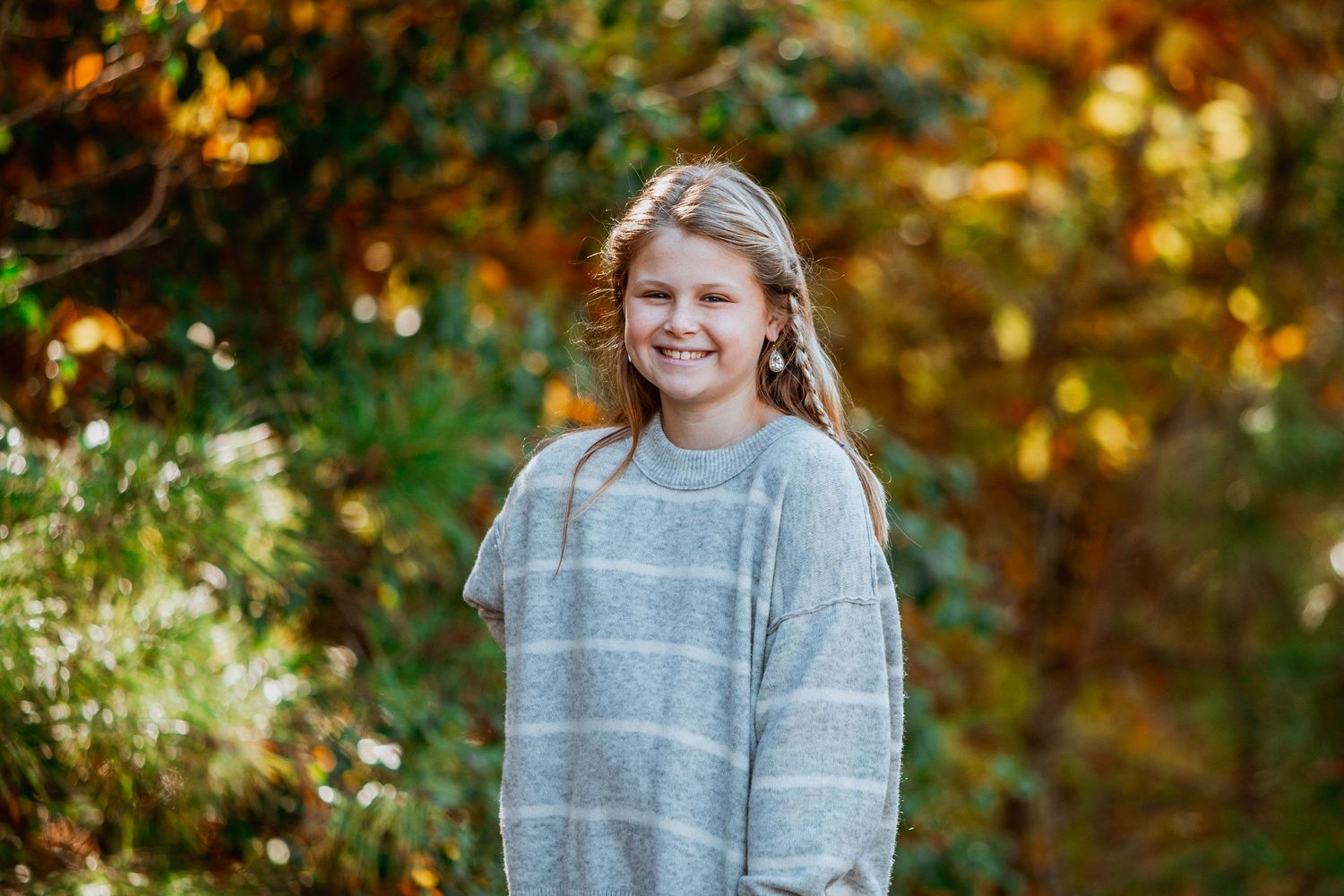 Smiling young girl in a gray striped sweater posing outdoors among colorful autumn foliage.