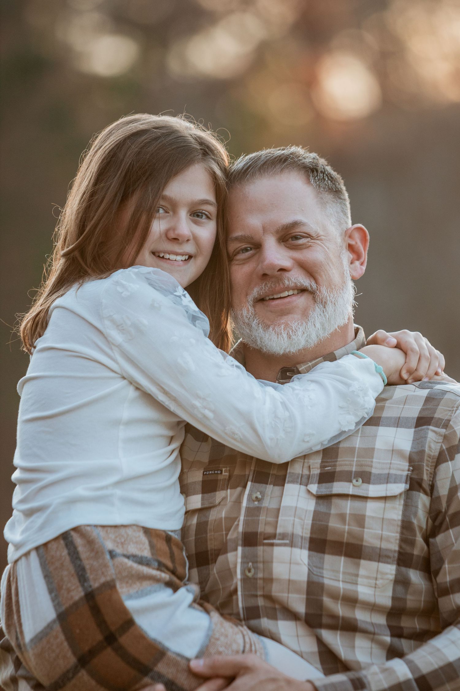Smiling girl hugging bearded father from behind in warm outdoor autumn light.