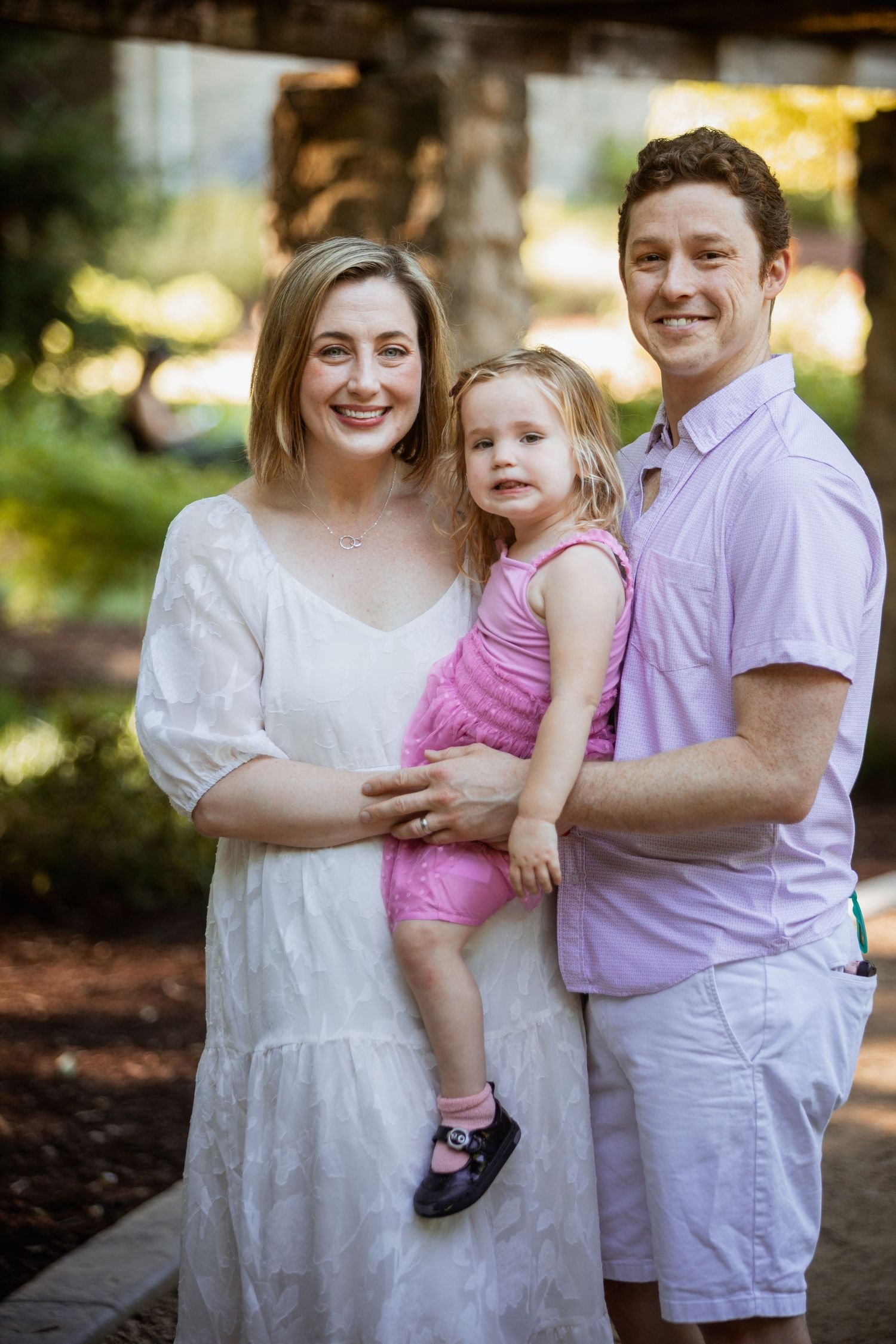 Happy family portrait with mom in white dress, dad in lavender shirt, holding toddler girl in pink outfit.