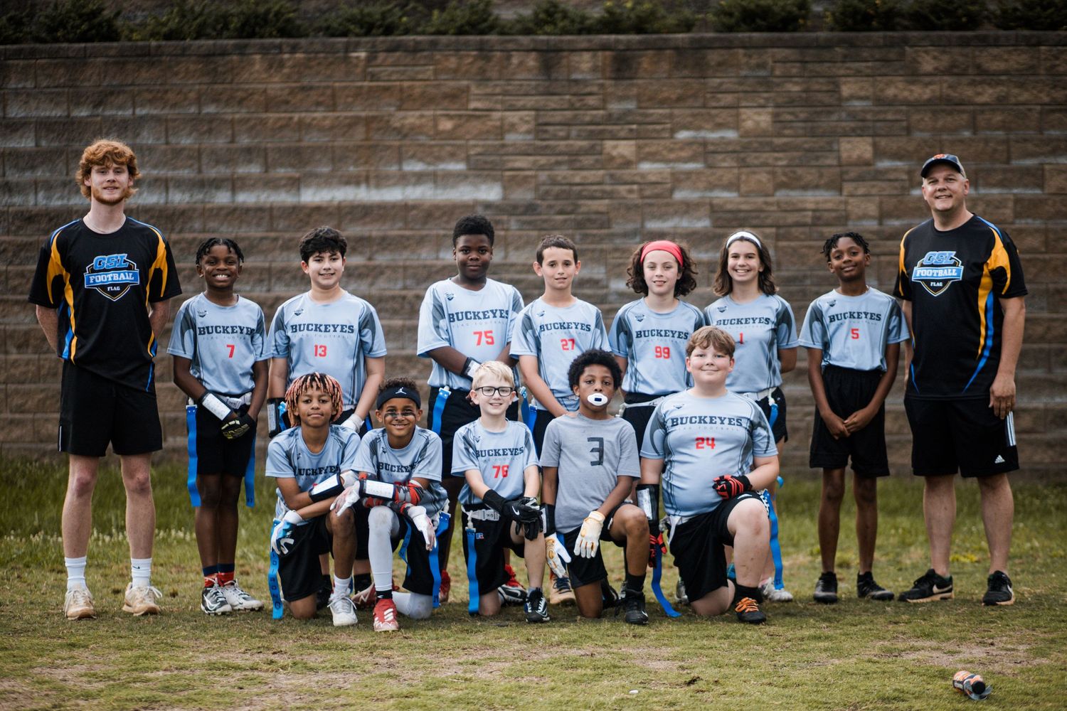 Youth flag football team in gray uniforms posing with two coaches against a brick wall.