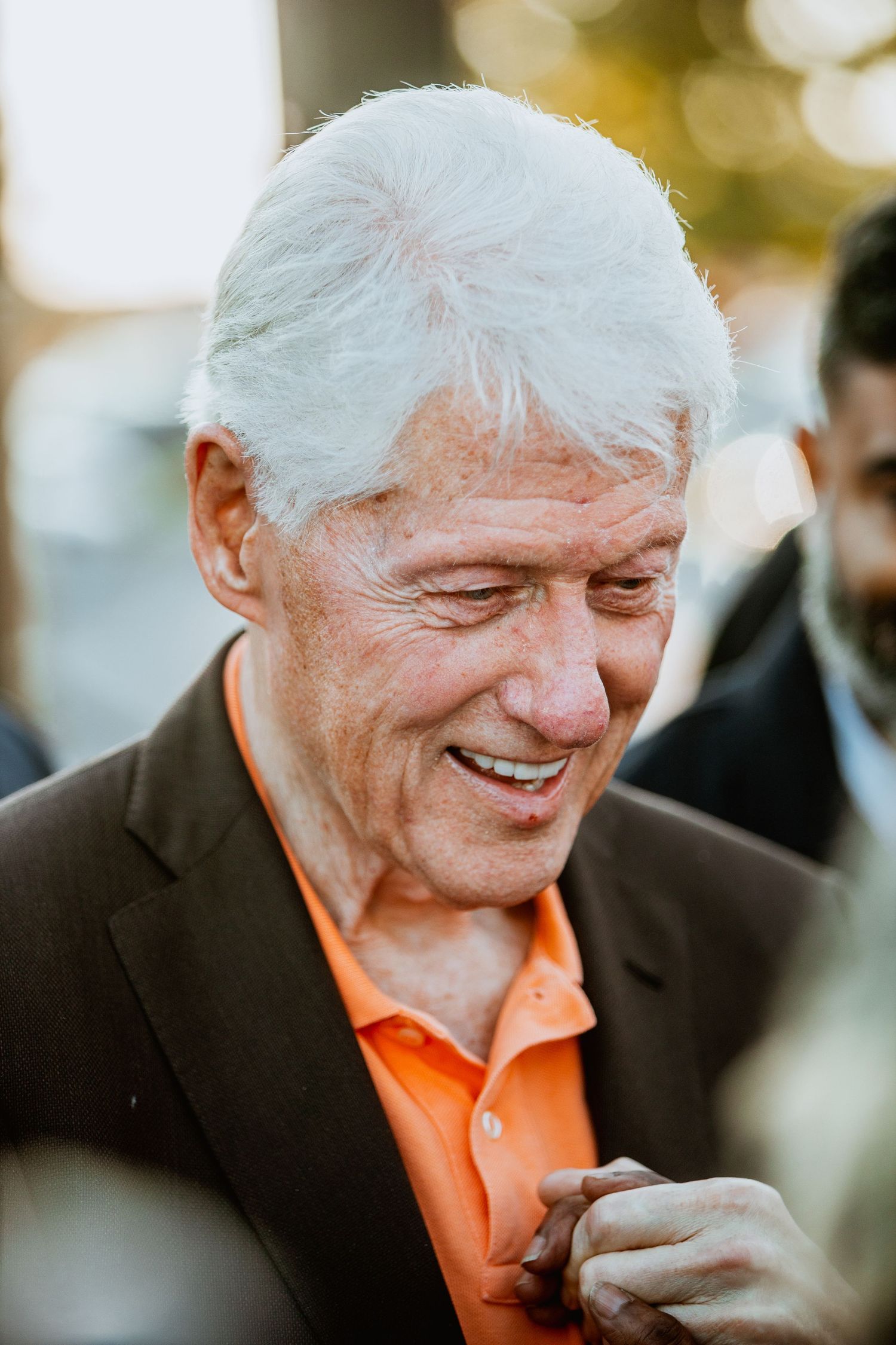 Smiling elderly man with white hair wearing an orange polo shirt and dark blazer outdoors.