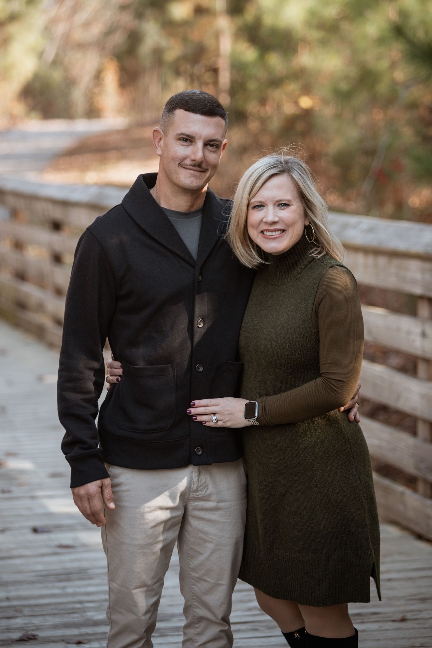 Smiling couple posing together on a wooden boardwalk in an autumn forest setting.
