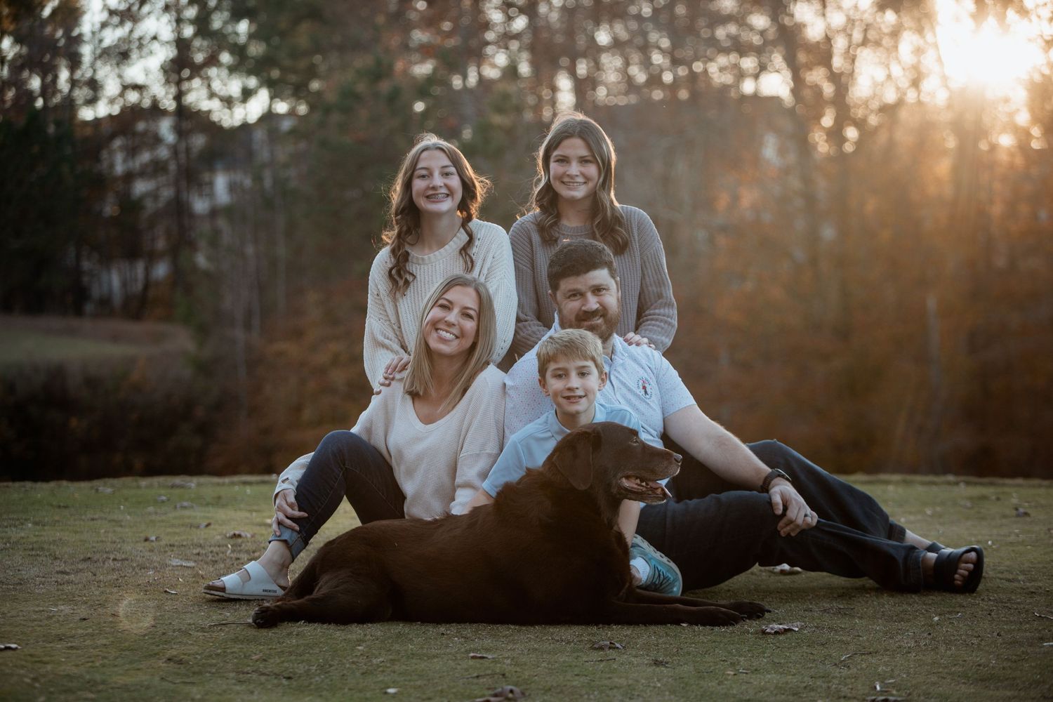 Family of five with a chocolate Labrador sitting together on grass during an outdoor fall photo session.