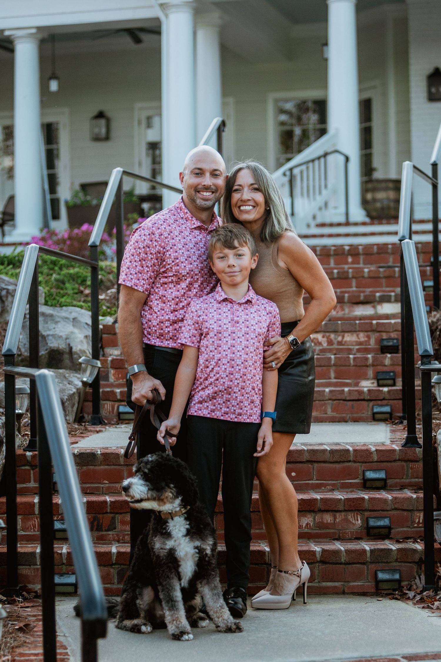 Family of three in matching pink polo shirts poses with their dog on brick steps outside a white columned home.