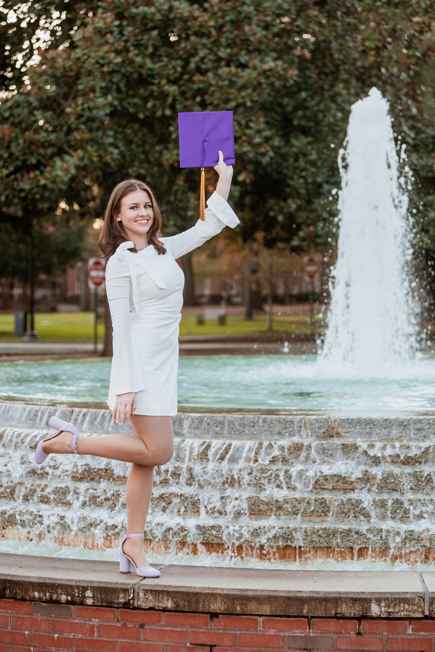 ECU graduate in white dress raises purple mortarboard cap triumphantly beside campus water fountain.