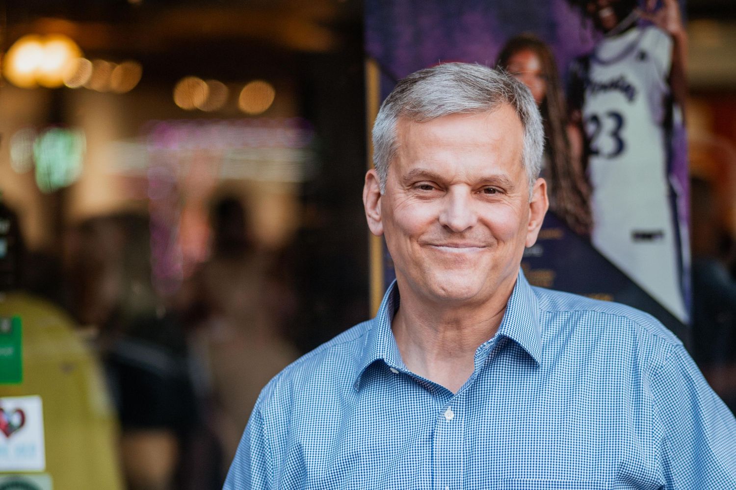 Smiling middle-aged man with gray hair wearing a blue button-up shirt standing in front of a sports store.