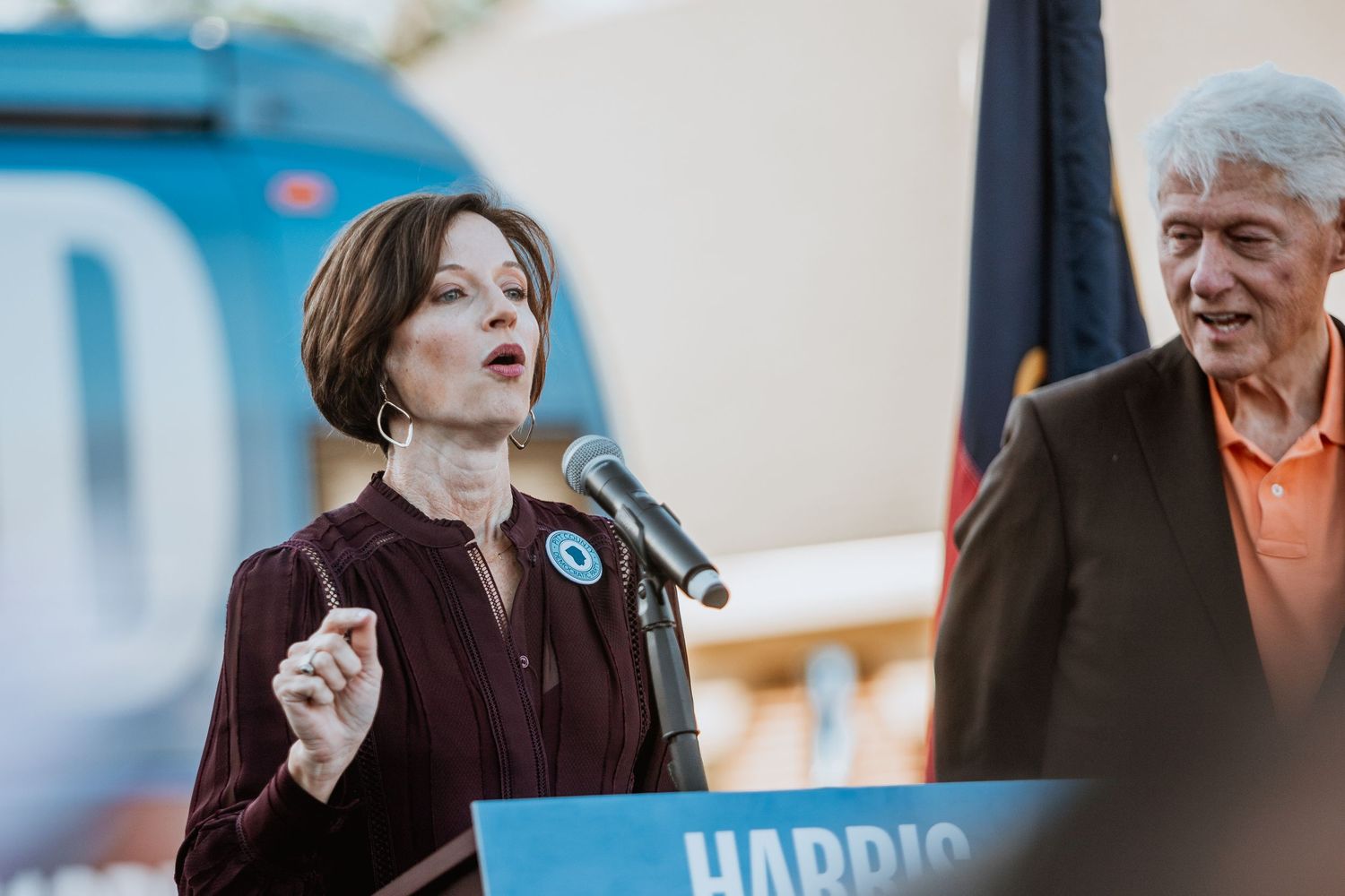 Woman speaks at Harris-Walz podium while older man in orange shirt listens at outdoor political rally.
