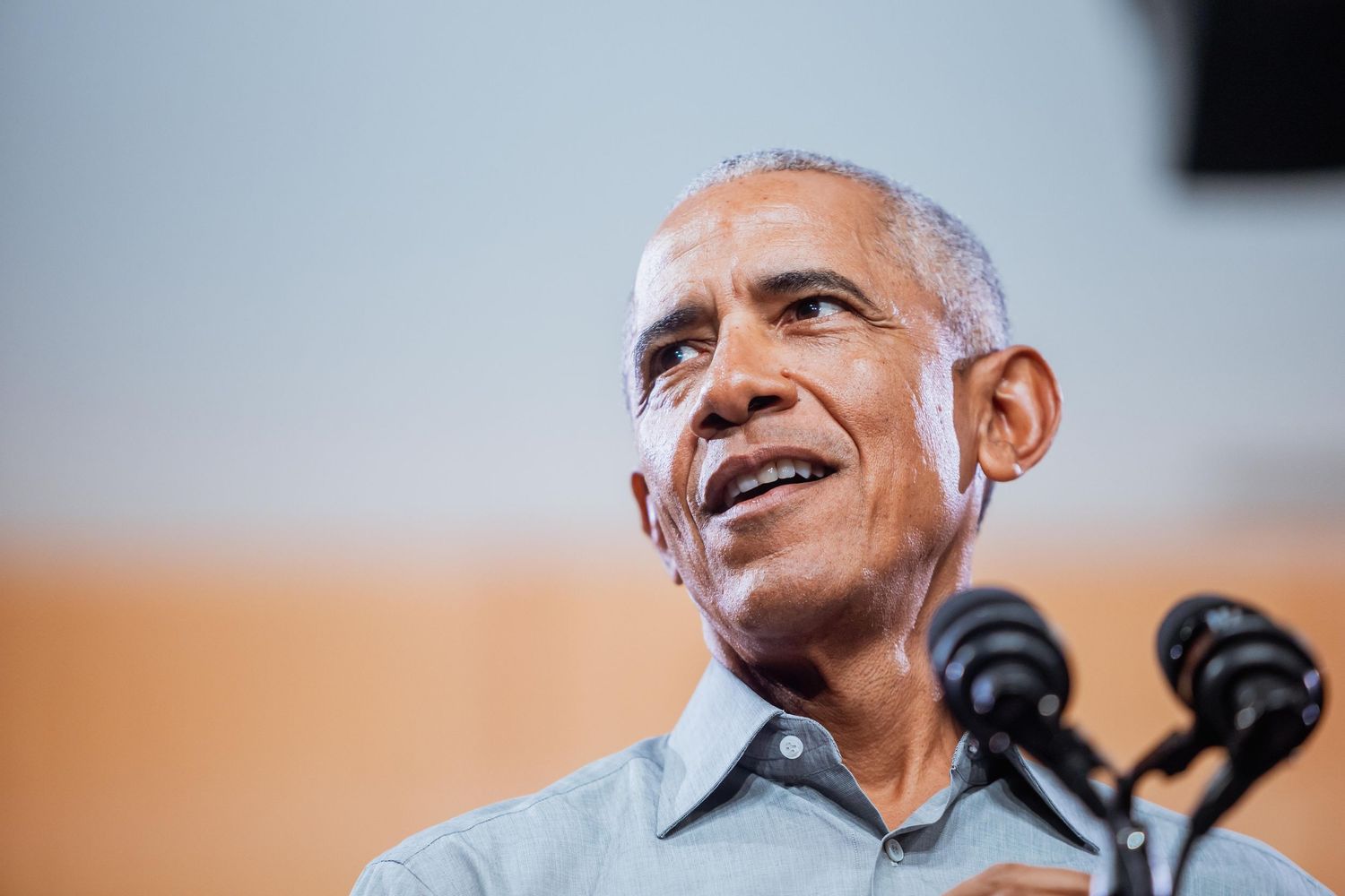 A man in a light blue shirt smiles while speaking at a dual-microphone podium.