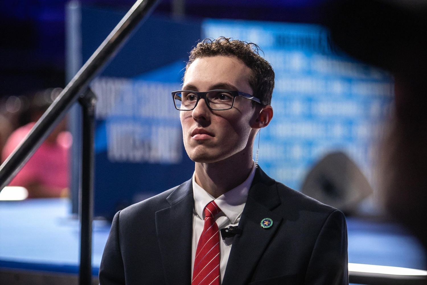 A young man in a suit and glasses stands attentively at a political event.