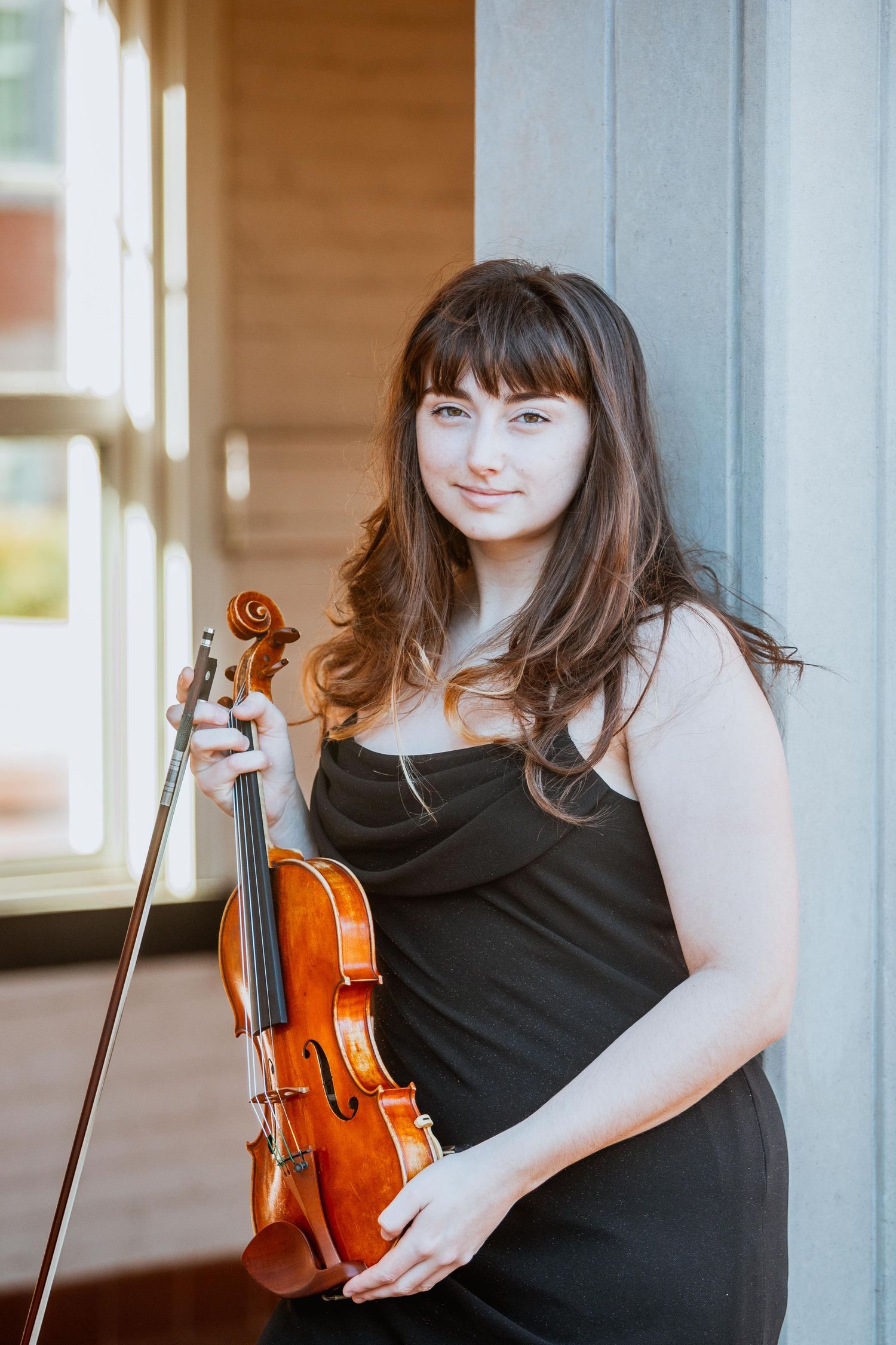Young woman with dark hair holding violin and bow in black dress indoors.