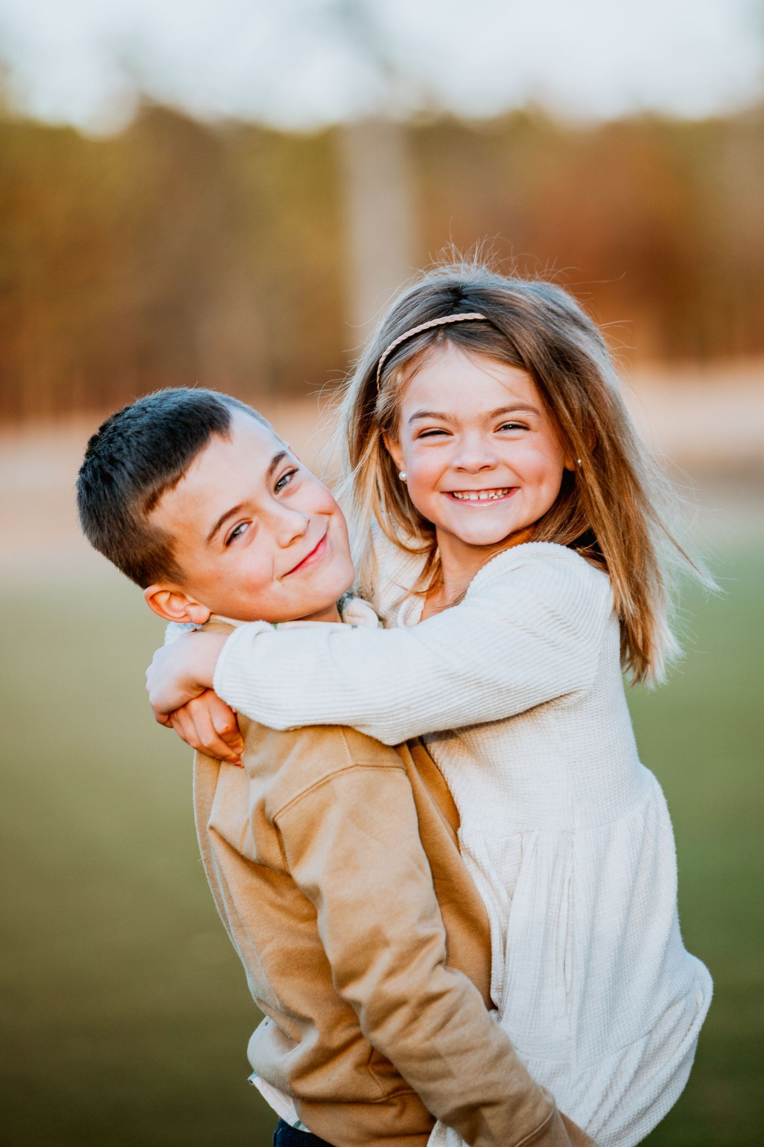 Two happy children hugging outdoors, girl smiling brightly while boy gives her a piggyback in golden afternoon light.