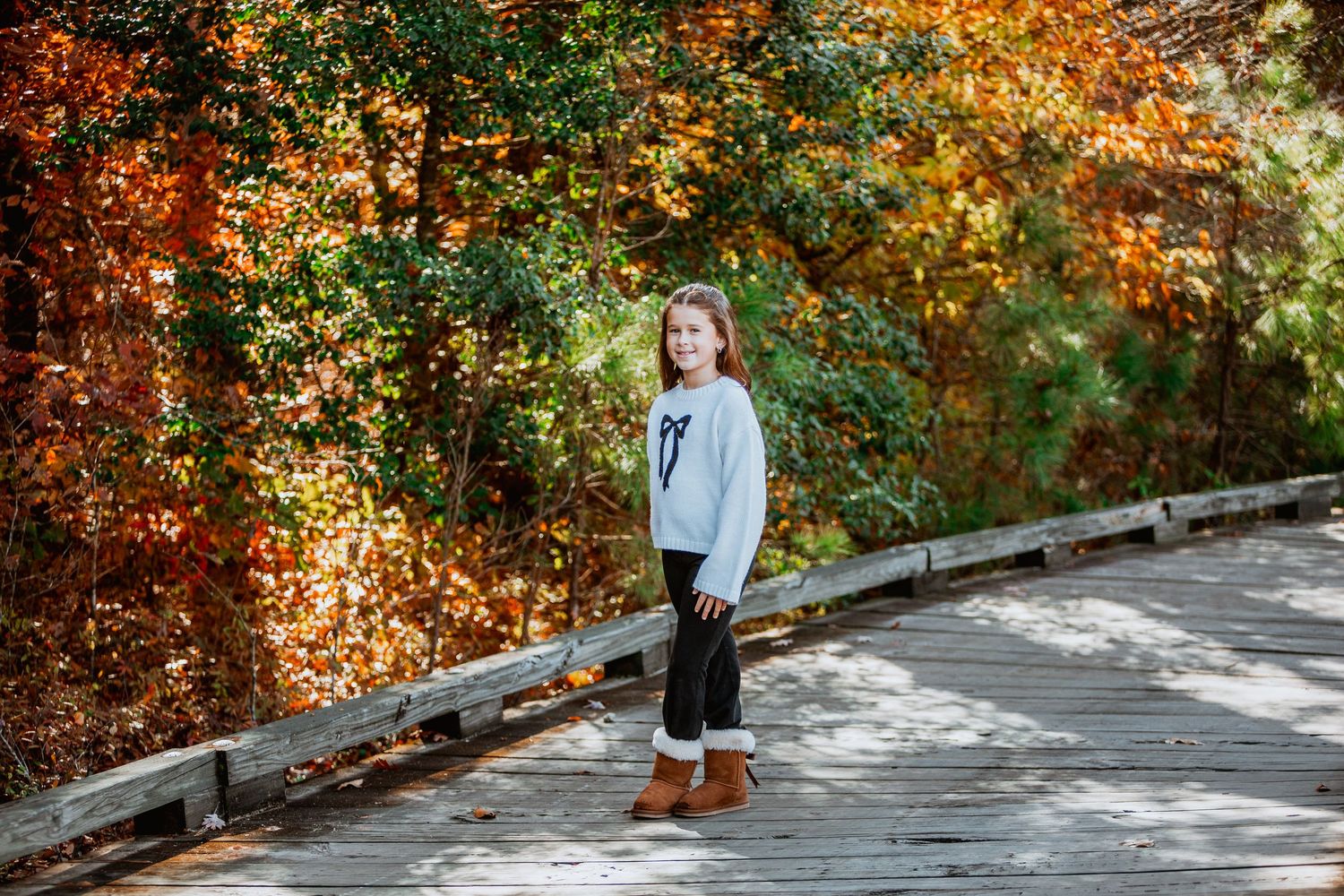 Girl in white bow sweater and brown boots standing on wooden bridge surrounded by fall foliage.
