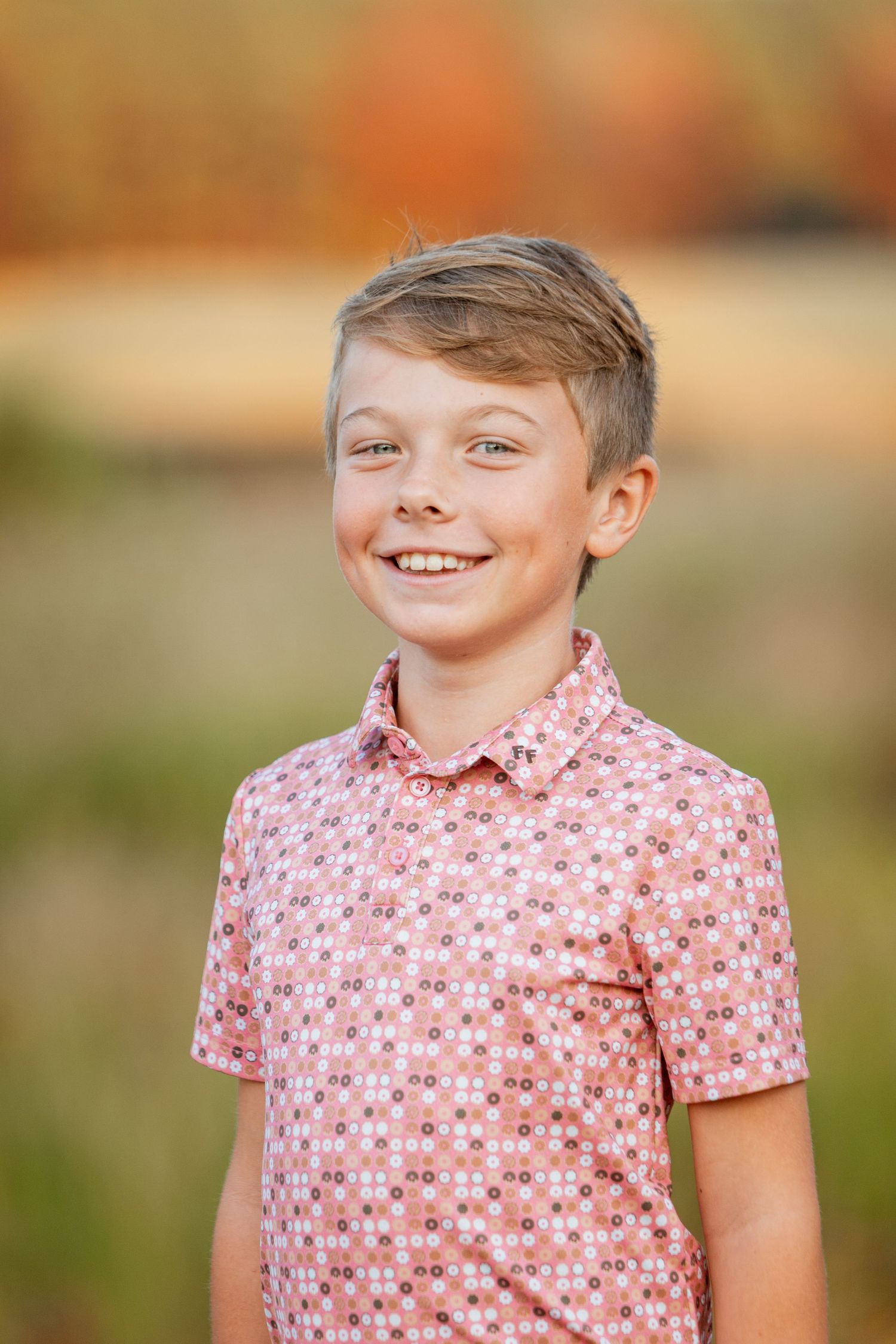 Smiling young boy in pink floral shirt poses for individual portrait with fall foliage background.