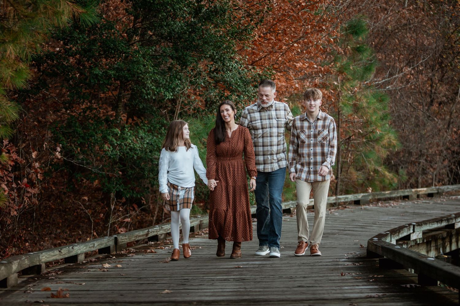 Family of four walking hand-in-hand on a wooden boardwalk with fall trees behind.