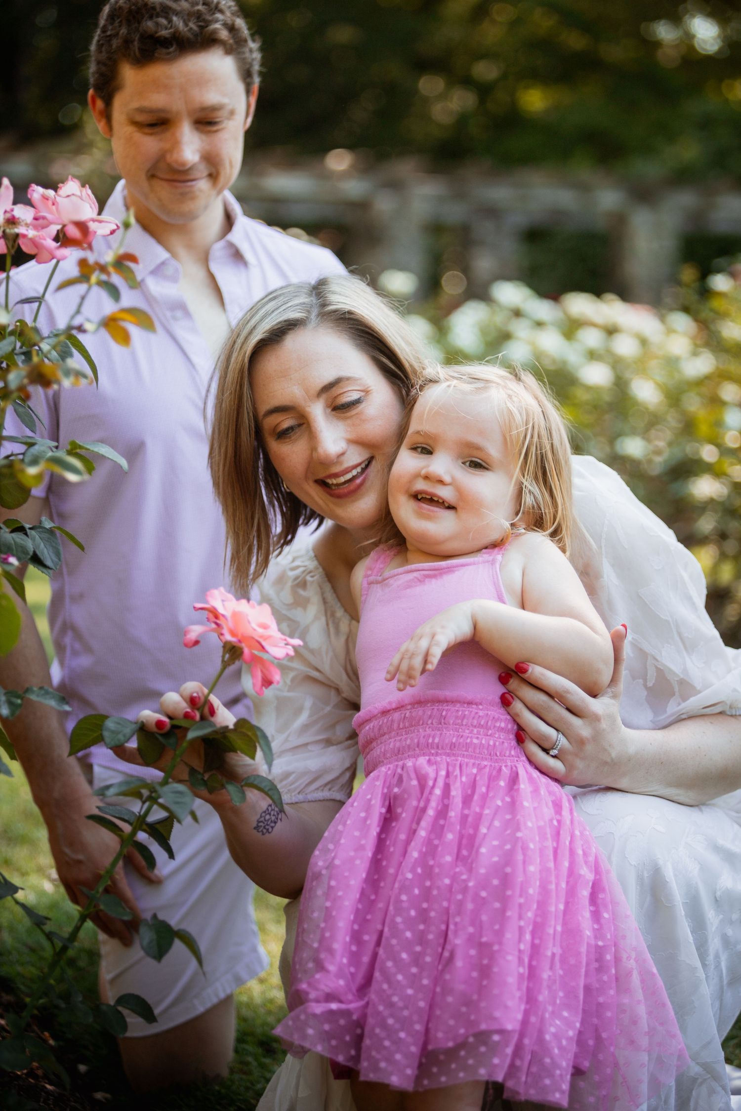Family of three smiling outdoors near pink roses, mother hugging toddler girl in pink dress with father beside them.