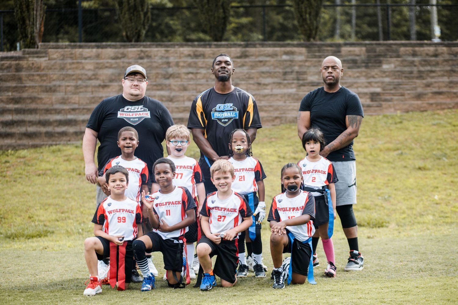 Youth football team poses with three coaches on a grass field wearing red and white uniforms.