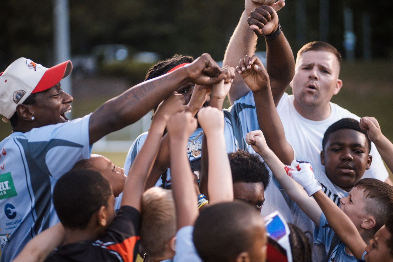 Coaches and youth football players gather in an enthusiastic team huddle on a grass field.