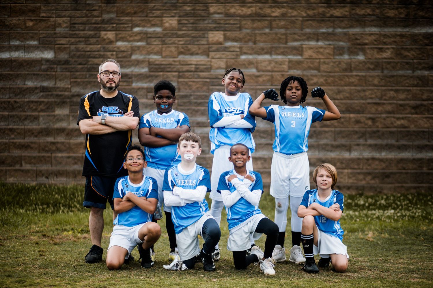 Youth flag football team in blue uniforms posing with coach against a brick wall outdoors.
