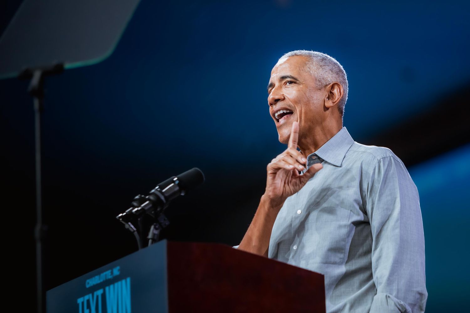 A cheerful man points upward while speaking at a wooden podium on stage.