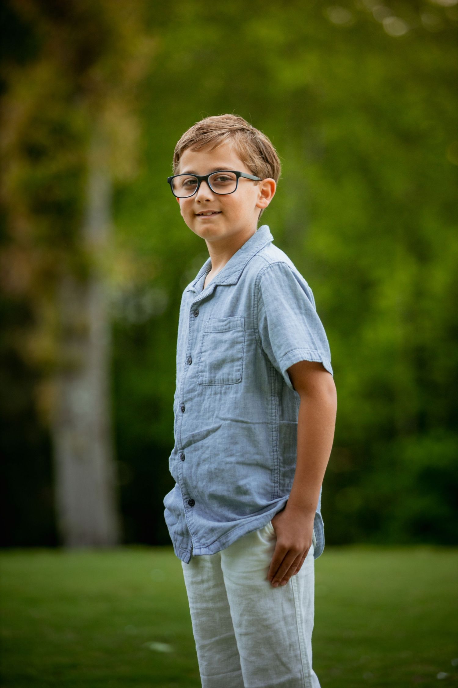 Young boy in glasses and blue shirt smiling confidently outdoors in a green park setting.