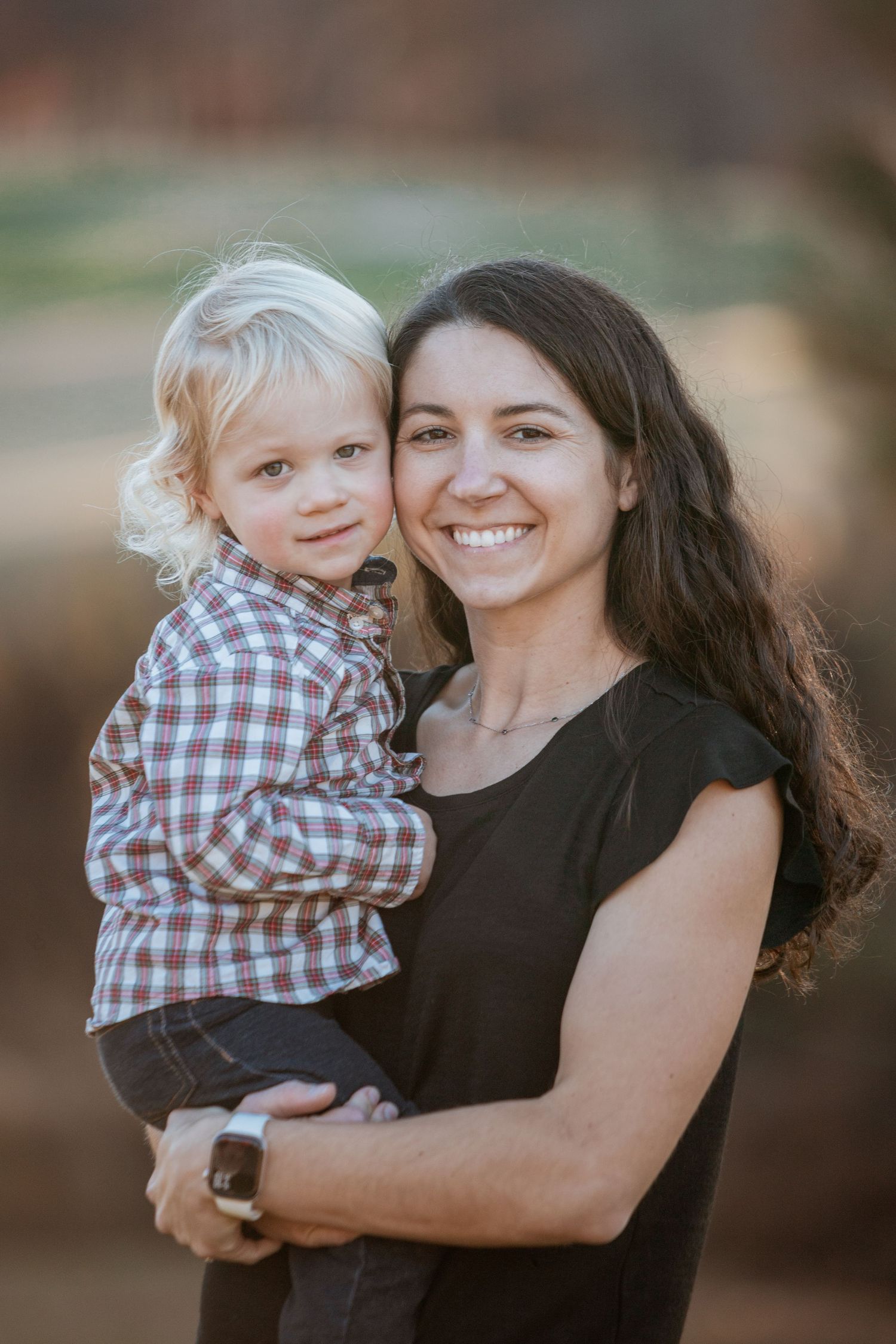 A smiling dark-haired mother holds her young blonde curly-haired son outdoors in warm evening light.
