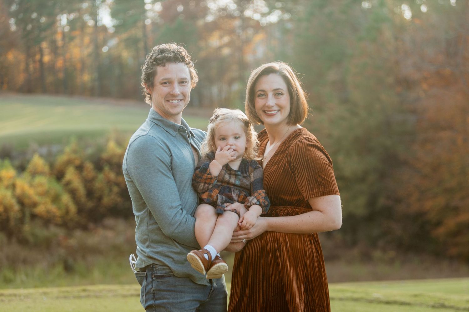 Smiling family of three with dad holding toddler daughter stands in golden autumn field at sunset.