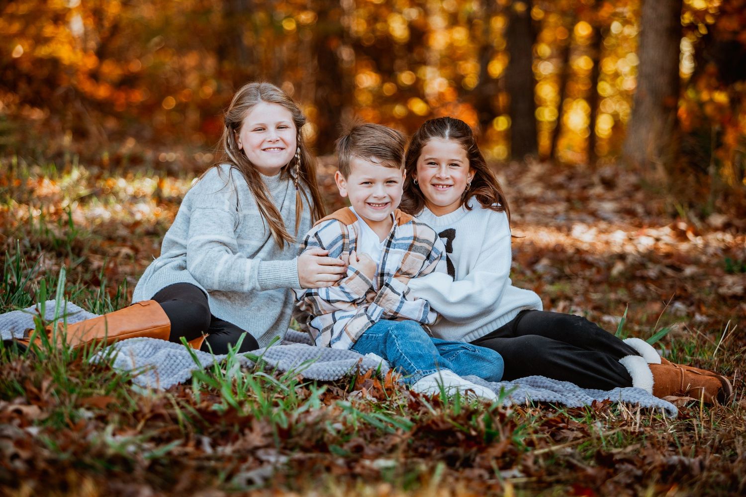Three happy siblings posing together on a blanket surrounded by fall foliage in the woods.