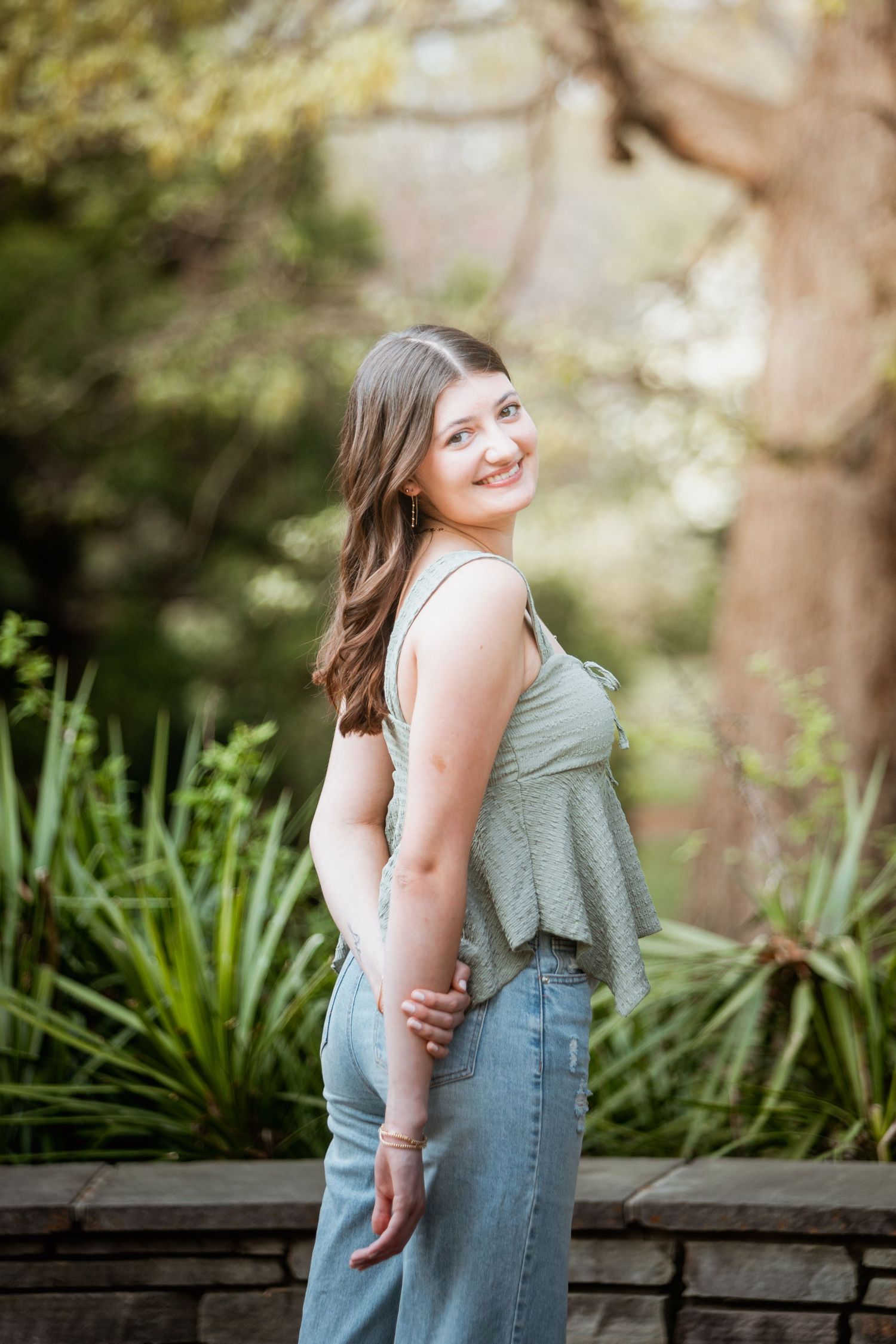 Smiling young woman in sage green top and jeans posing in a lush garden setting.