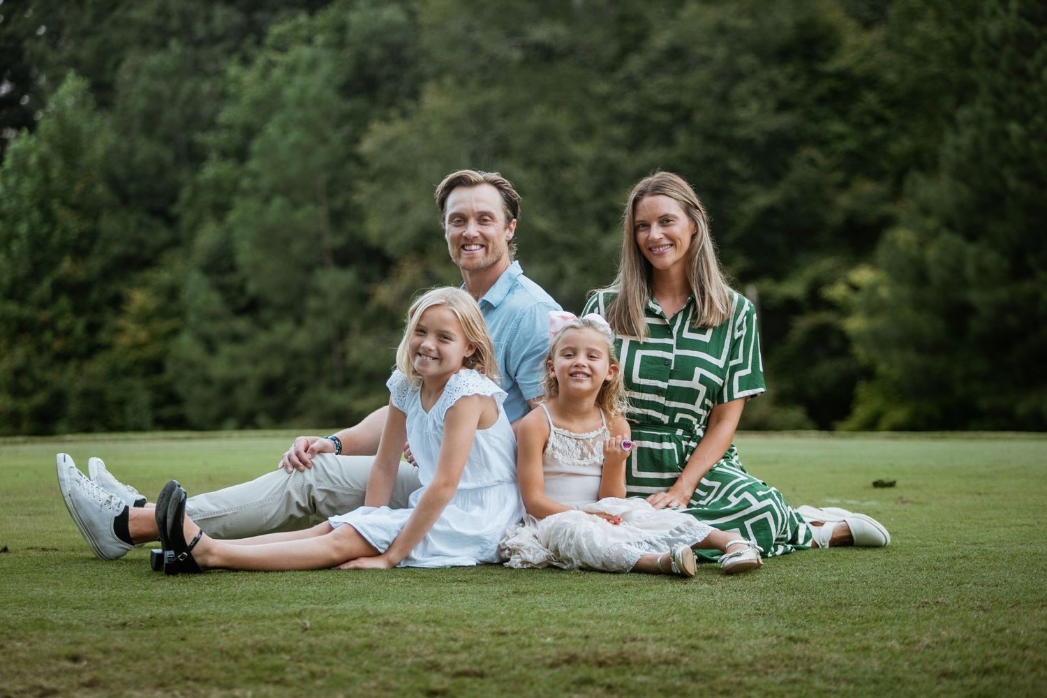 Happy family of four sitting together on green grass with trees behind them.