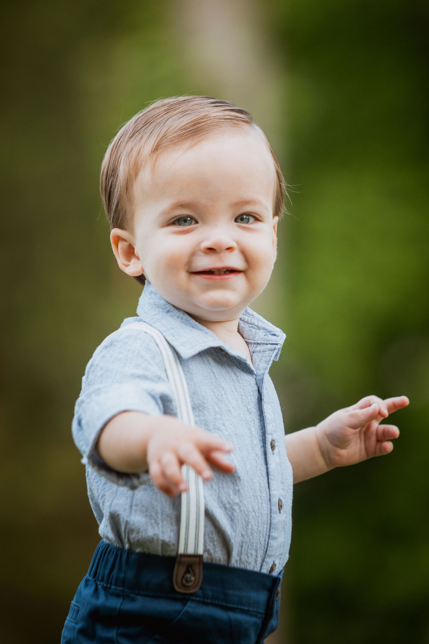 Smiling toddler boy in blue shirt and suspenders reaching out in a lush green outdoor setting.
