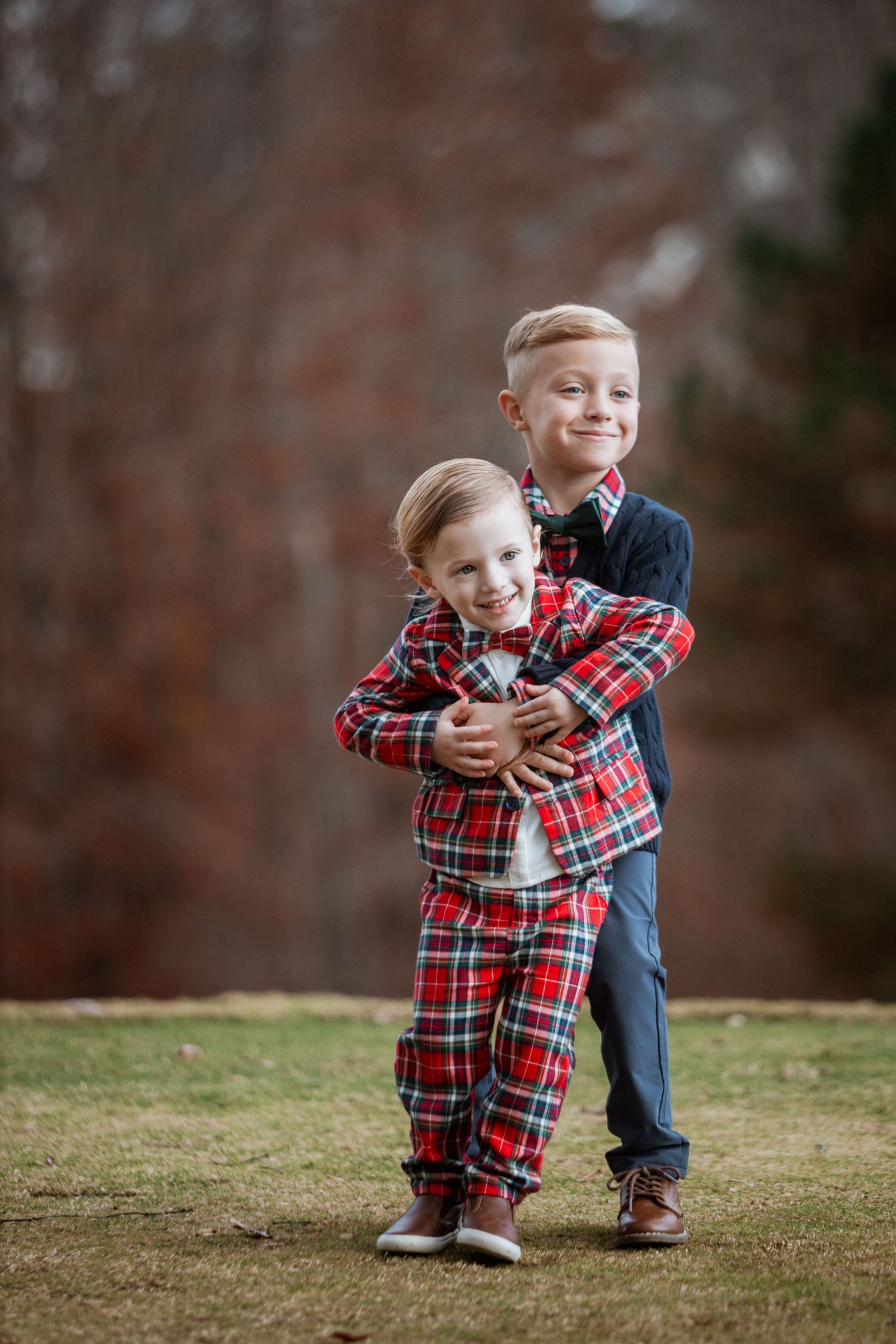 Older boy in navy hugs younger toddler in red plaid outfit on a grassy outdoor setting.