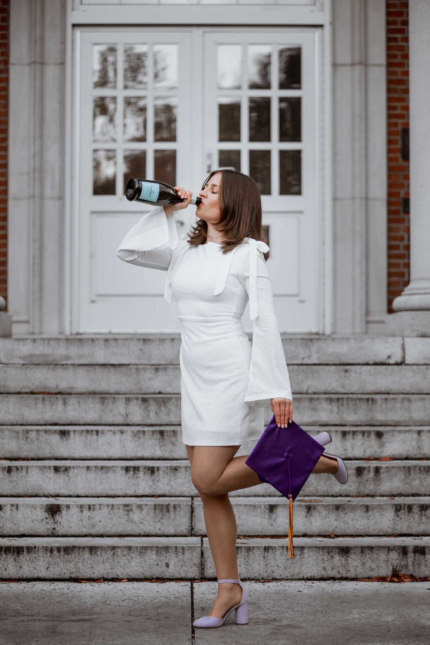Woman in white dress celebrating graduation, drinking champagne on steps while holding purple cap.