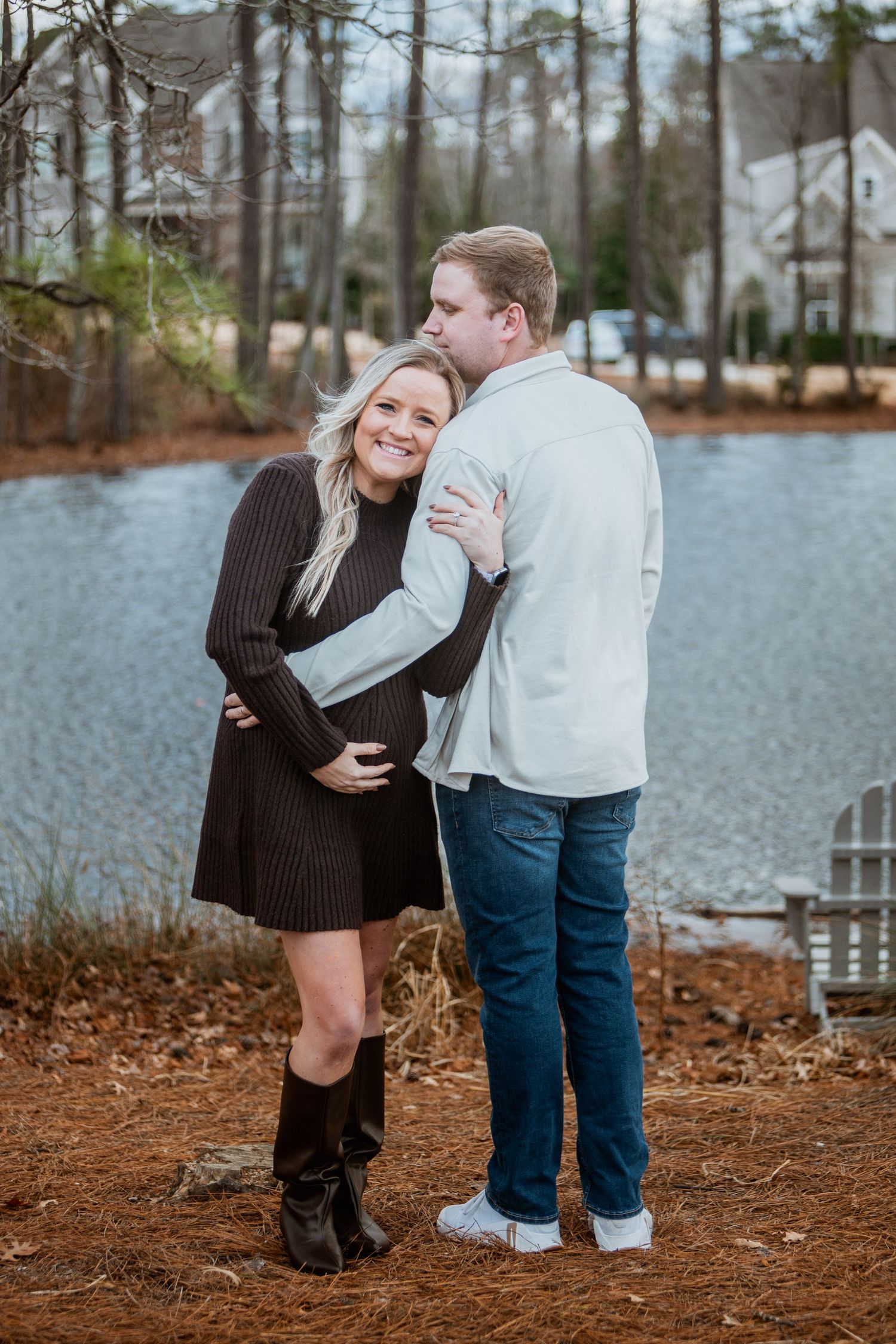 Pregnant woman in brown dress and boots embraces her partner near a lake in winter.