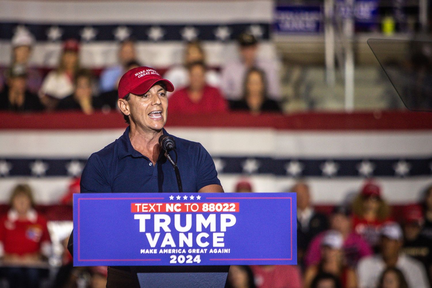 A man in a red Trump cap speaks at a Trump-Vance 2024 campaign rally podium in North Carolina.