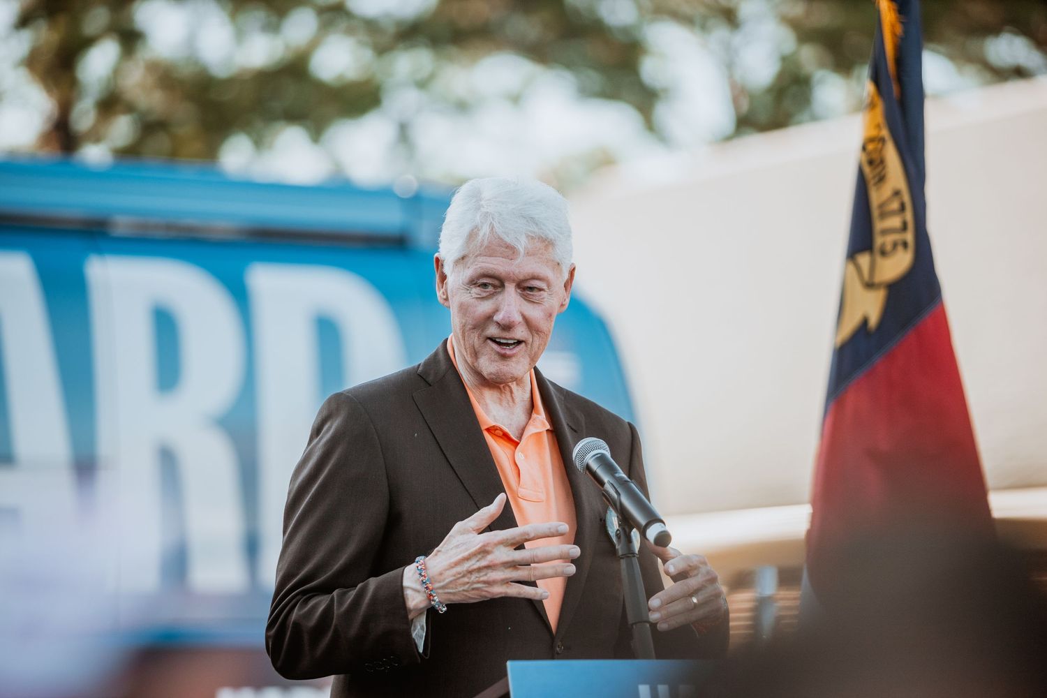 Senior man in orange shirt and blazer speaks at outdoor Harris Walz political rally.