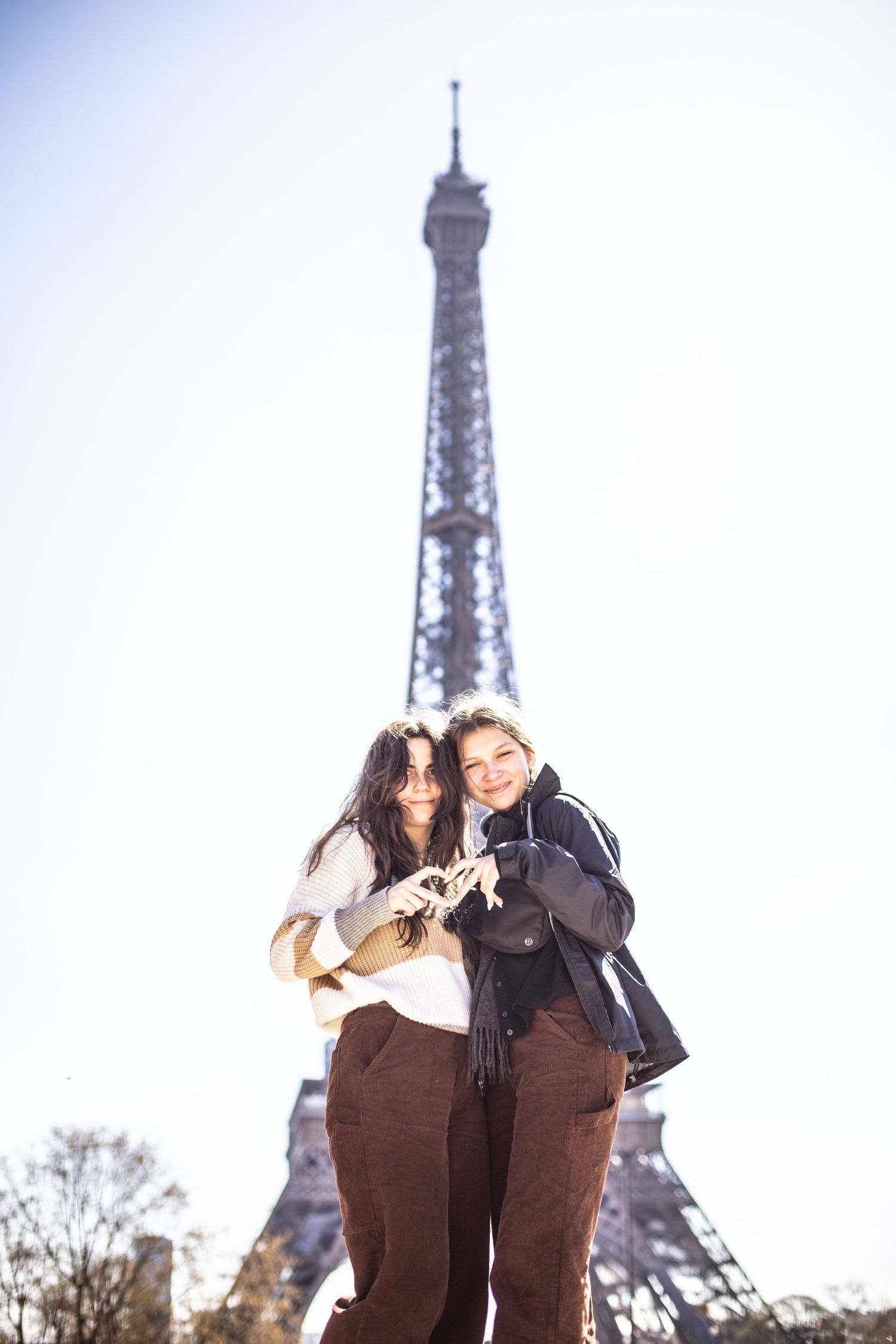 Two friends making a heart shape with their hands in front of the Eiffel Tower on a sunny day.