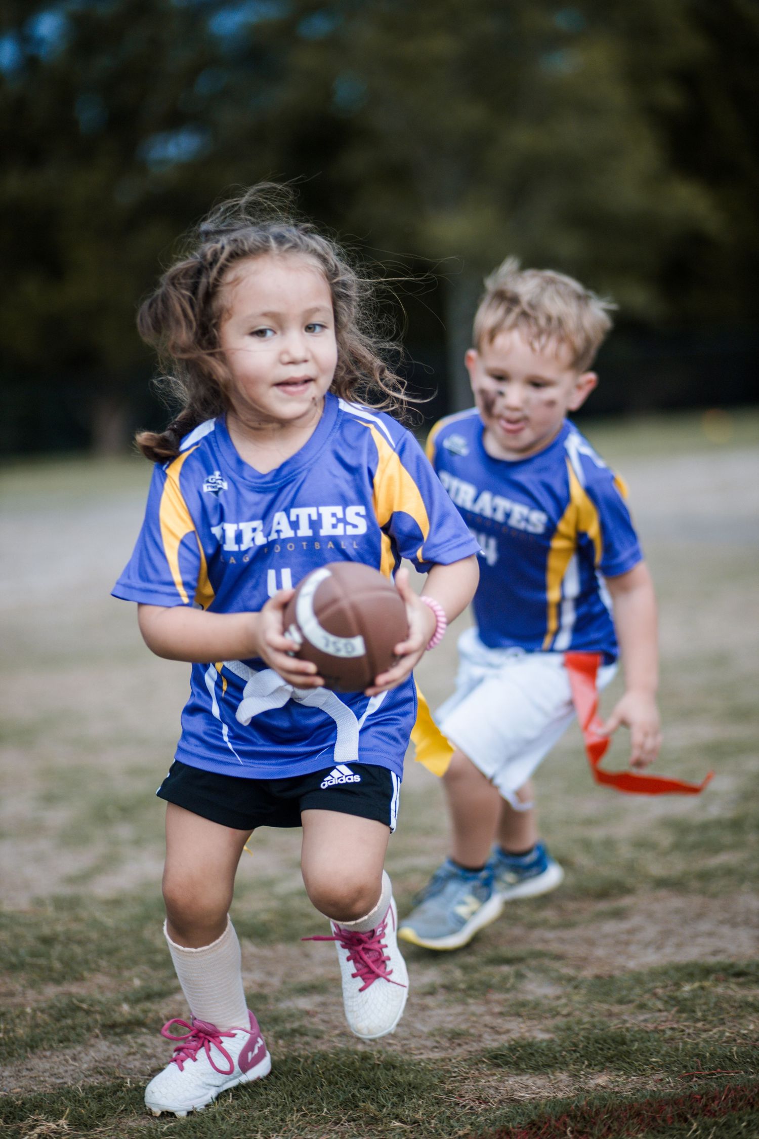 Girl in Pirates jersey runs with football as boy chases during youth flag football.