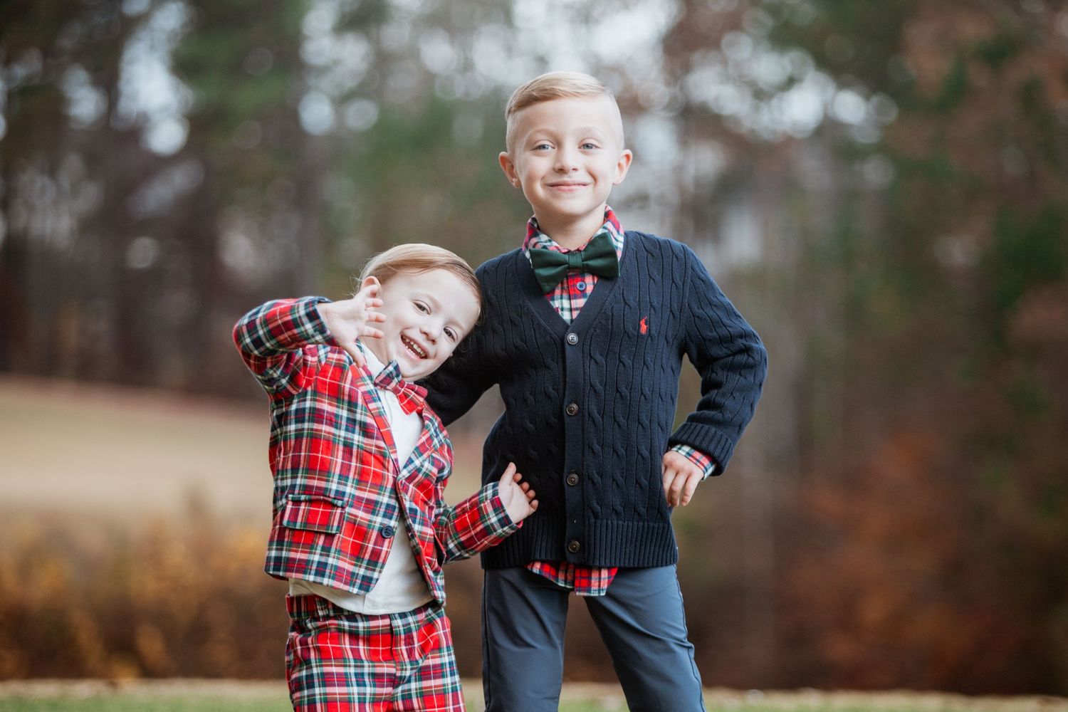 Two young boys in festive holiday attire smile and pose playfully outdoors in an autumn setting.