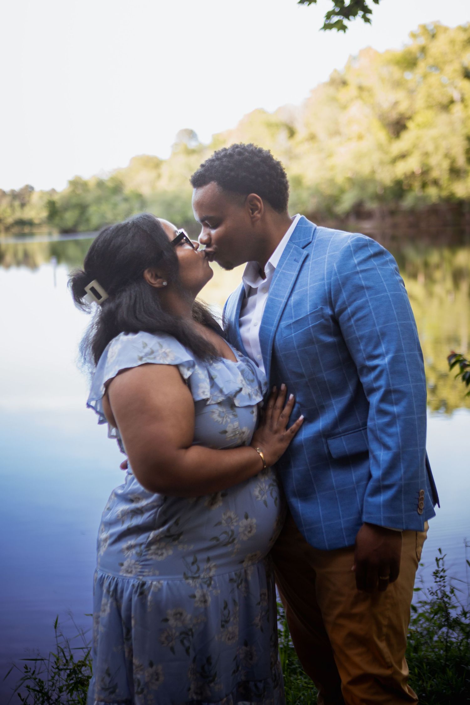 Couple sharing a romantic kiss near a serene lake, woman in floral blue dress and man in blue blazer.