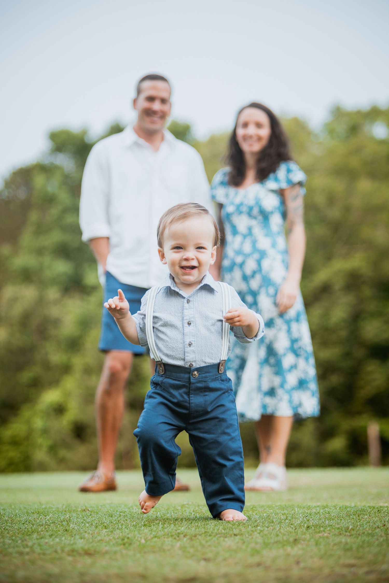 Joyful toddler boy running toward camera while parents smile proudly in a green outdoor setting.