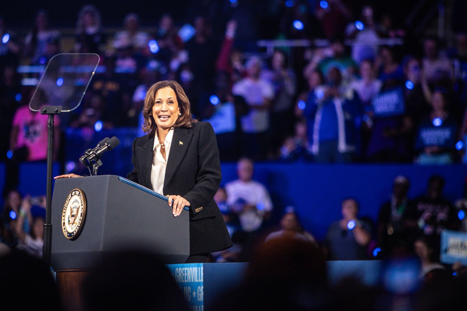 A smiling female politician stands at a podium during a Harris-Walz campaign rally before a large audience.