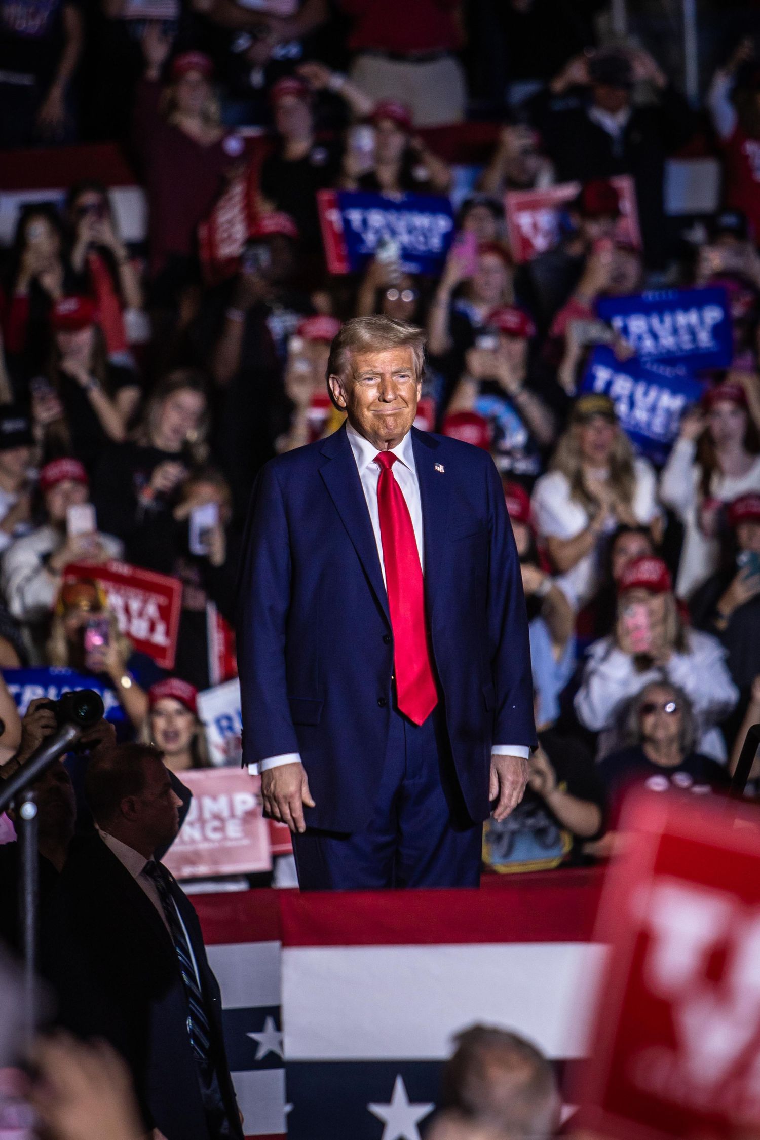 Donald Trump smiles at a packed campaign rally with enthusiastic supporters holding Trump Vance 2024 signs.