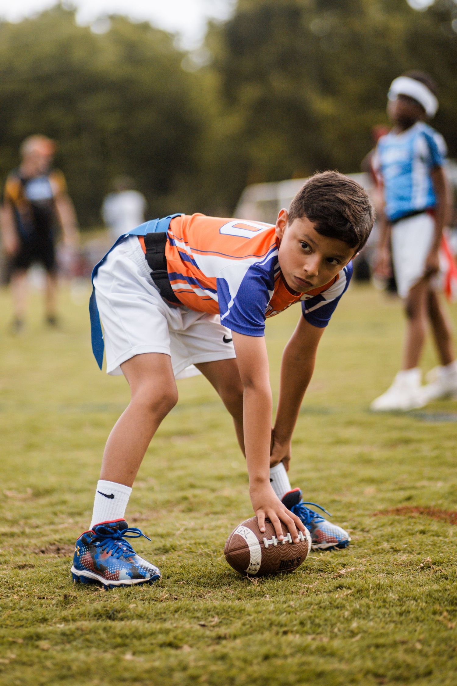Youth football player in orange and blue jersey crouching to snap the ball on field.