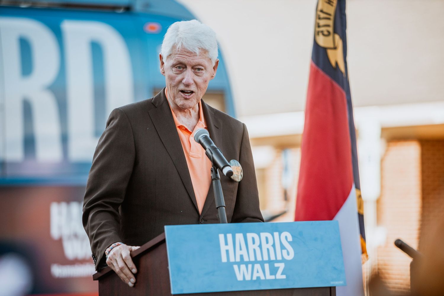 Man in brown suit speaks at Harris Walz campaign podium with North Carolina flag in background.